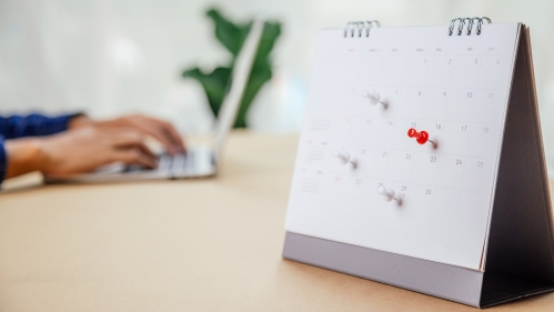 Calendar with push pins on select dates with person working on computer in background