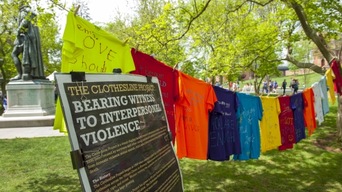 Photo of Rutgers Clothesline Project: Sign reads "Bearing Witness to Interpersonal Violence" and t-shirts of various colors hang from a clothesline between two trees, the t-shirts have words on them