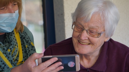 Two people looking at a phone; one wearing a medical mask to cover mouth and nose; the other an older woman smiling at the phone