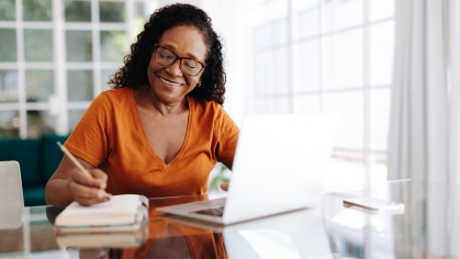 Older woman smiling while writing on notepad and working on computer