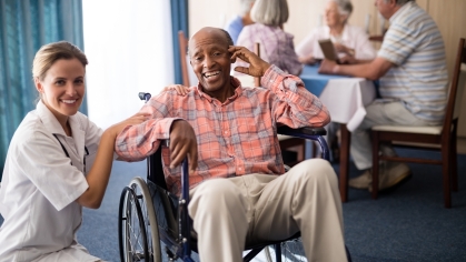 stock image of a female presenting health practitioner kneeling down next to an elderly male presenting person of color using a wheelchair. both are smiling at the camera
