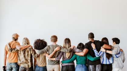 Group of nine young adults standing in a line with their arms around each other's backs, the camera's point of view is behind the people in the photo