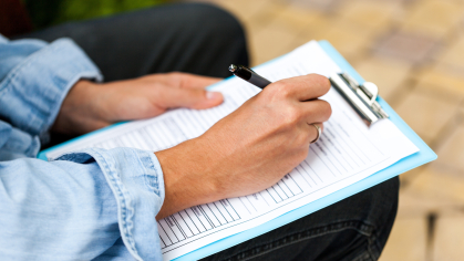 Person sitting holding a clipboard and pen and completing a paper survey. Image of only hands and legs.
