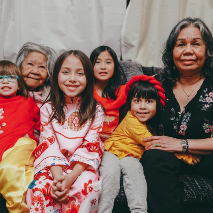 Image of family sitting on a couch looking at camera; multi generational family members from young children to elders
