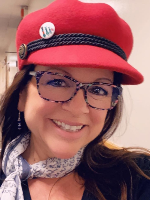 Close cropped photo of a woman with glasses and brown hair smiling and looking at the camera; she is wearing a red hat