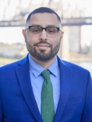 Brown Latino with a short beard, wearing glasses, a blue suit, blue shirt, green site, standing in front of the Queensboro Bridge