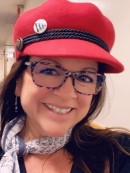 Close cropped photo of a woman with glasses and brown hair smiling and looking at the camera; she is wearing a red hat