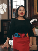 Image of a woman standing in a well appointed room and smiling at the camera; she's leaning her hand on a table and wearing a black and red outfit