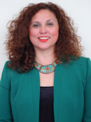 Close cropped photo of a woman with curly red, brown hair; she is smiling at the camera and wearing a decorative necklace and a green jacket