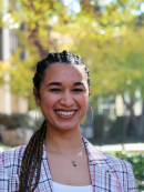 photo of a woman in the foreground standing outside and smiling at the camera; she has long dark hard that is braided at the top and in a ponytail to the side