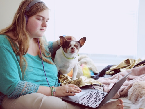 White woman sitting on a bed with headphones in and using her laptop with her small dog sitting next to her