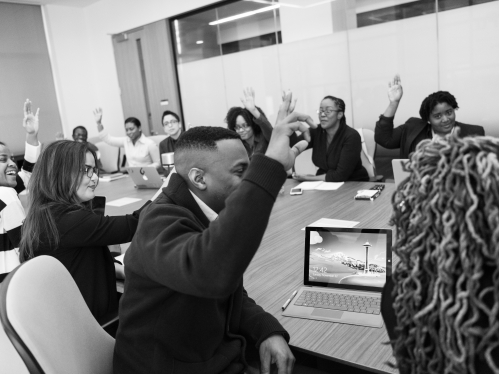 Black and white photo with a group of people in professional clothing sitting around a conference table with most of them raising their hands
