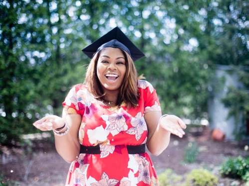 Black female in a red and white floral dress smiling and facing the camera with a graduation cap on