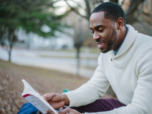 Black male in a white sweater sitting outside reading a paperback book