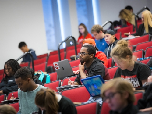 Large lecture hall with various students working at their seats. One student with their laptop in the Center of the image but not at the front