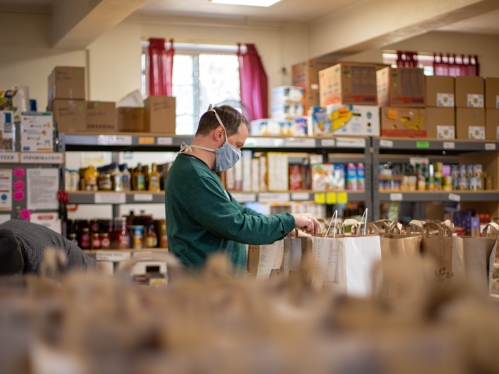 Large room with shelves of food and a person at the center with a medical mask on to cover their mouth and nose while they pack groceries and organize food