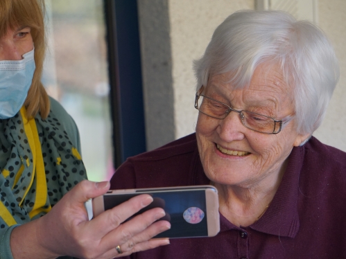 Two people looking at a phone; one wearing a medical mask to cover mouth and nose; the other an older woman smiling at the phone