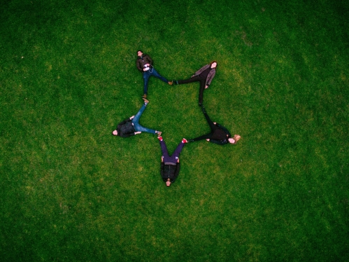 zoomed out photo taken from the sky of people laying in the grass with their feet touching to make a star pattern
