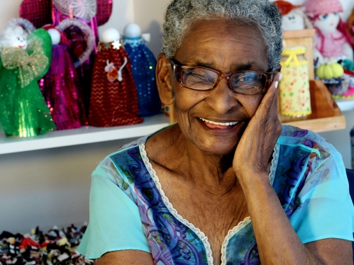 older black woman looking at the camera in a bright blue top with black glasses on; smiling and holding her hand to her face