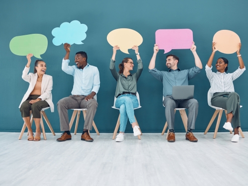 Stock image of a group of people sitting on chairs smiling and looking at each other. Each are holding up a blank word bubble