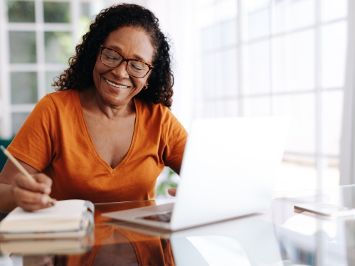 Older woman smiling while writing on notepad and working on computer